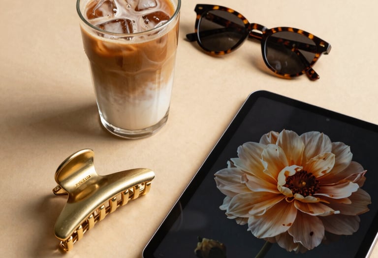 An aesthetic flat lay of a chic morning setup on a warm beige table. A glass of iced latte, a gold hair claw clip, a pair of stylish tortoise-shell sunglasses, and a digital tablet showing a floral screensaver. North American / US lifestyle.
