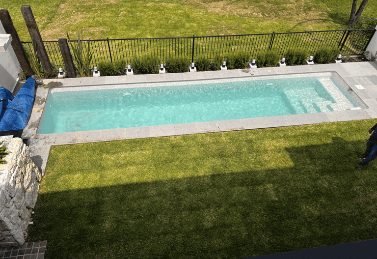 Alberca rectangular con escalones, rodeada de jardín verde y barda de seguridad metálica | KARÄNA