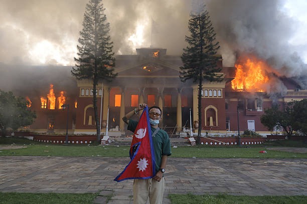A man holds a Nepal flag in front of a large historic building engulfed in flames and thick smoke.