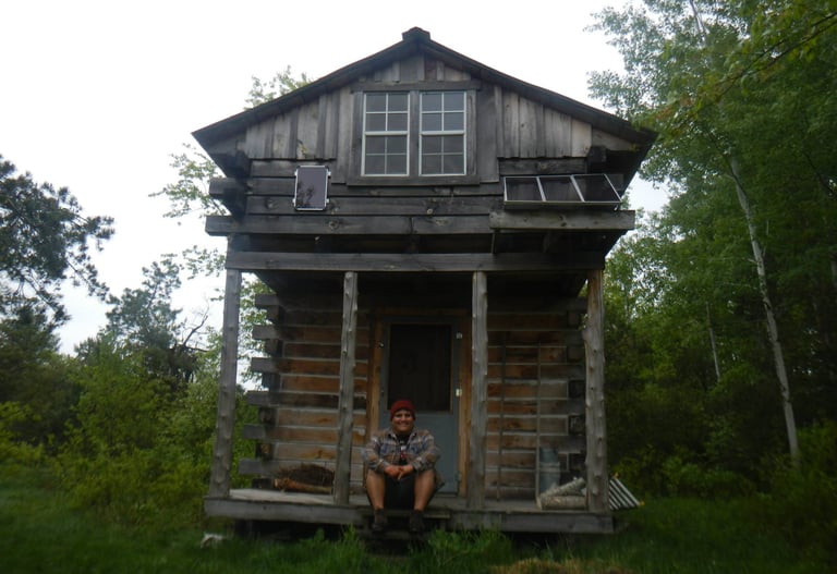 a smiling person sits on the front porch of a rustic wood cabin