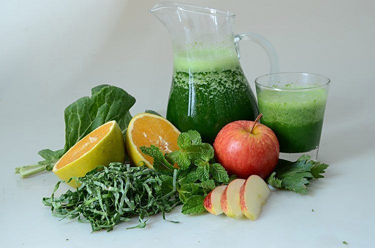 Jarra e copo de suco verde cercados por ingredientes frescos como laranja, maçã, couve, hortelã 