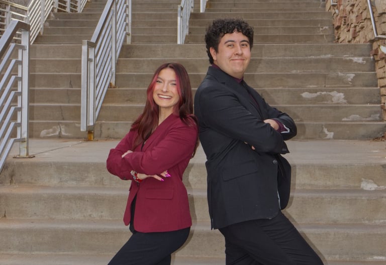 Victoria Quesada-Stoner and Ben Gregg standing back to back with hands folded, and stairs behind.