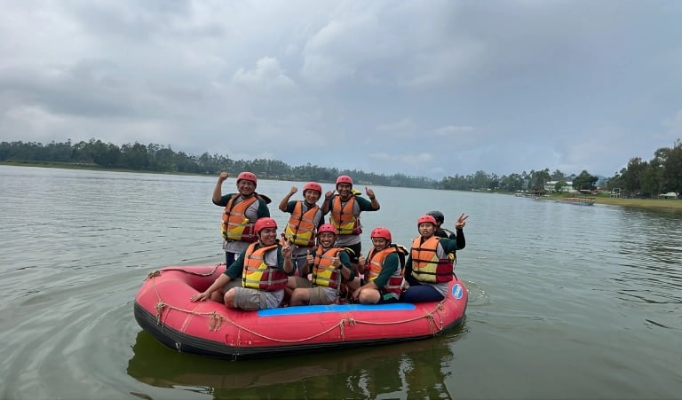 Delapan orang bersemangat di perahu karet merah melakukan rafting di Situ Cileunca Bandung, mengenakan jaket pelampung oranye