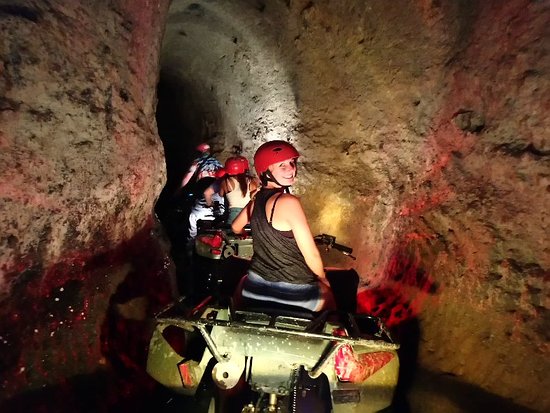 Adventurous tourists wearing red helmets ride ATVs through a narrow limestone underground cave tunnel.