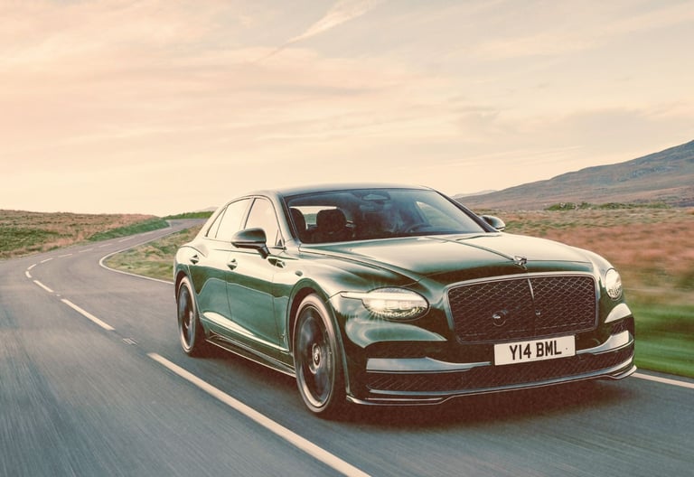 A dark green Bentley Flying Spur luxury sedan driving on a winding countryside road at sunset.