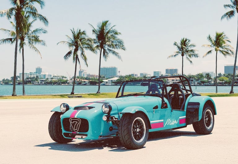 A blue Caterham Seven sportscar parked in Miami with palm trees and a city skyline background.