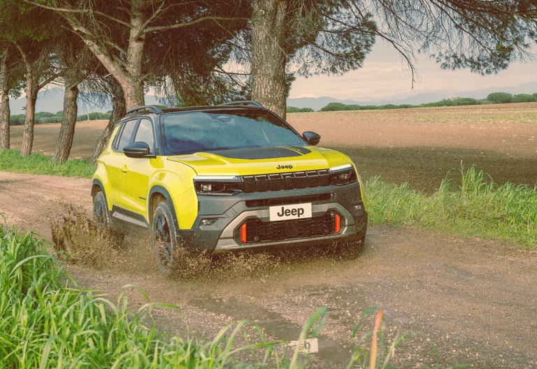 Yellow Jeep Avenger 4xe driving through a mud puddle on a dirt road lined with trees.