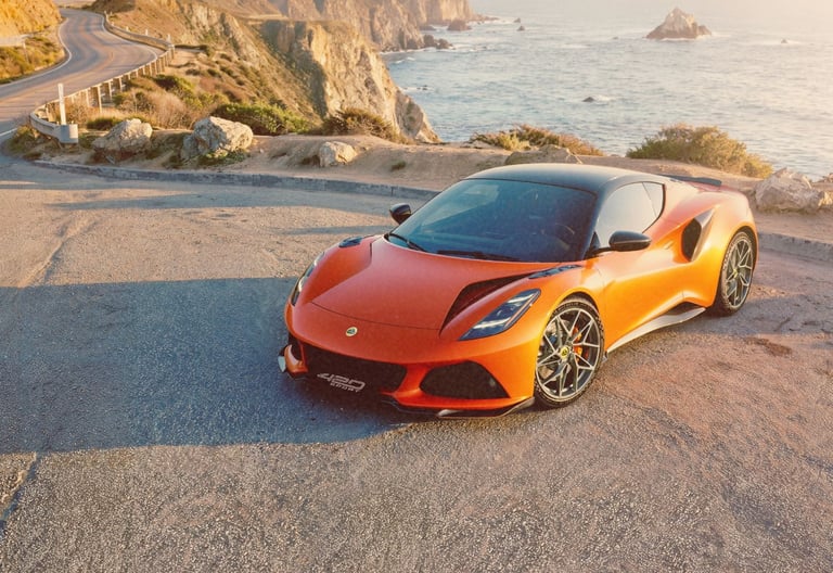 Orange Lotus Emira sports car parked on a scenic coastal cliff road overlooking the Pacific Ocean.