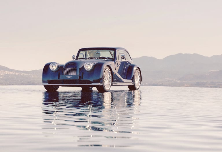A vintage-style dark blue Morgan sports car parked on a reflective surface with mountain views.