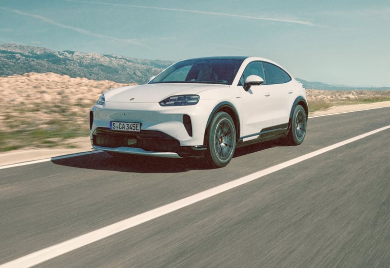 White Porsche Cayenne Electric driving on a coastal road with mountain scenery.