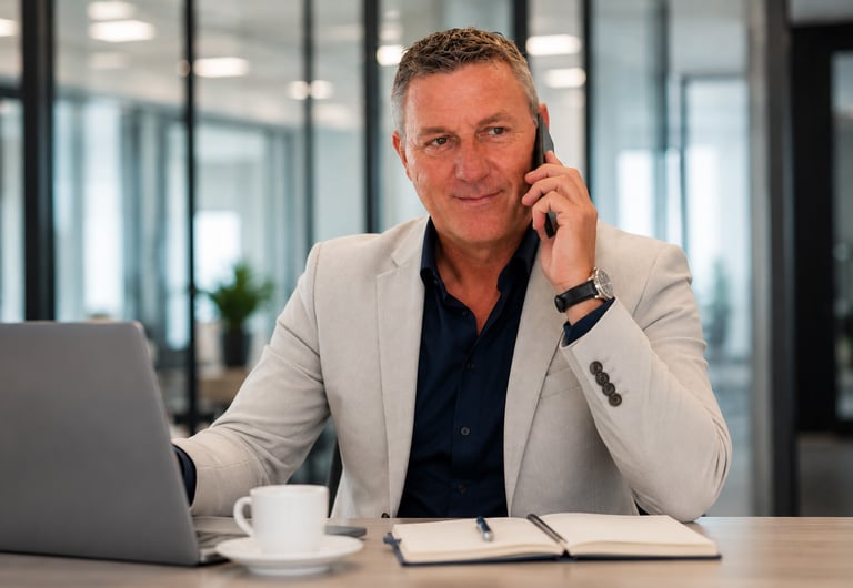 Professional businessman in a suit talking on a mobile phone while working on a laptop in a modern office.
