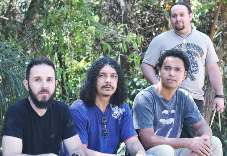 A four-member rock band with long hair and beards posing outdoors in front of green trees.