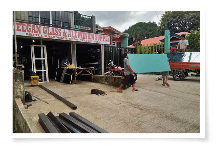 Two workers carrying a large panel outside a glass and aluminum supply store, with a dog resting on the pavement.