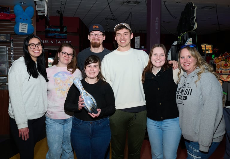 A group of smiling friends posing at a bowling alley while holding a decorated souvenir bowling pin.