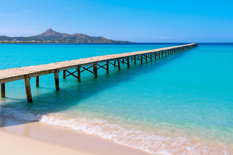 Playa de Muro en Mallorca con muelle de madera sobre aguas turquesas y montañas al fondo – Maiorca Unlocked.