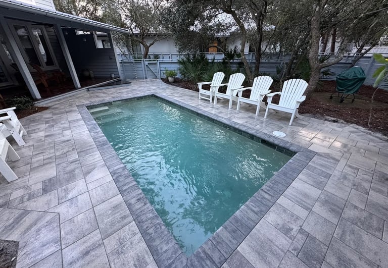 A rectangular backyard plunge pool with stone pavers and white Adirondack chairs in a lush patio setting.