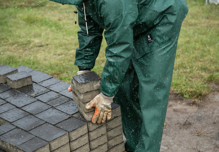 Worker in green rain gear stacking concrete paving stones for a patio landscaping project in the rain.