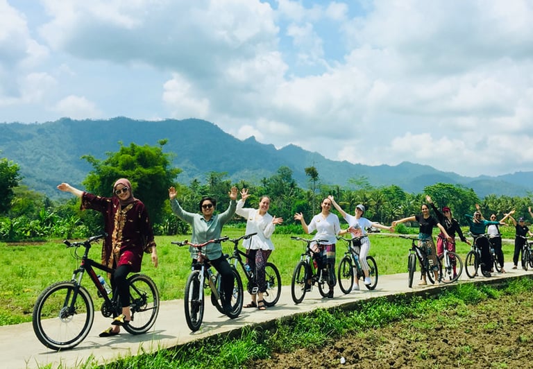 Wisatawan bersepeda santai menyusuri jalan pedesaan dengan pemandangan sawah dan Candi Borobudur di kejauhan