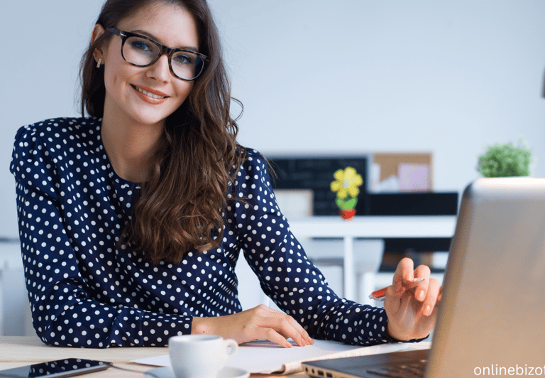 a woman in glasses and a polka dot shirt is smiling and holding a pen with a laptop on the desk
