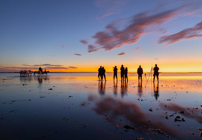 Photographers and horses silhouetted against a vibrant orange sunset on a reflective salt flat water surface.