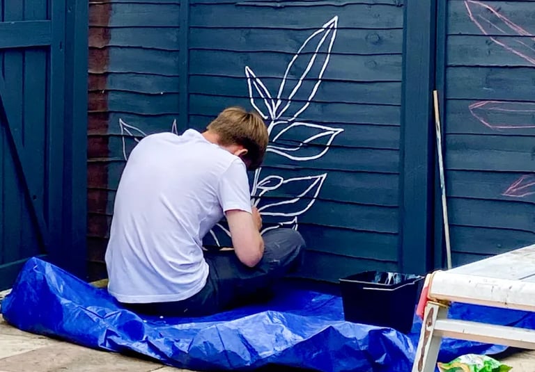 Muralist Byron sits on floor, painting graphic plant on grey wooden garden fence.