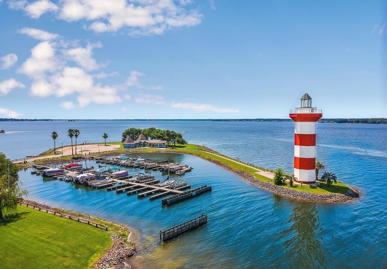 Aerial view of a red and white striped lighthouse at a boat marina on a sunny day with blue water.