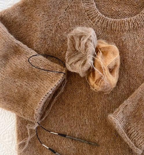 Unfinished beige hand knit sweater lying on a checked blanket with knitting needles and yarn.