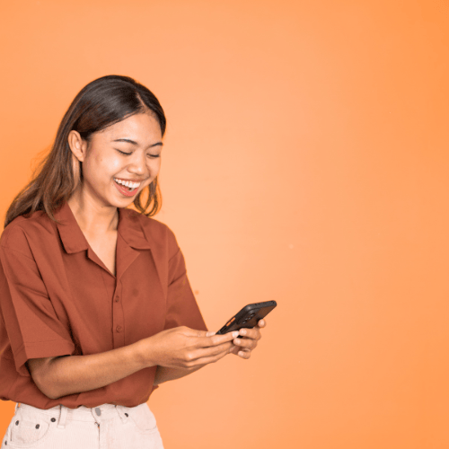 Smiling young woman holding a smartphone and laughing while texting on an orange background.