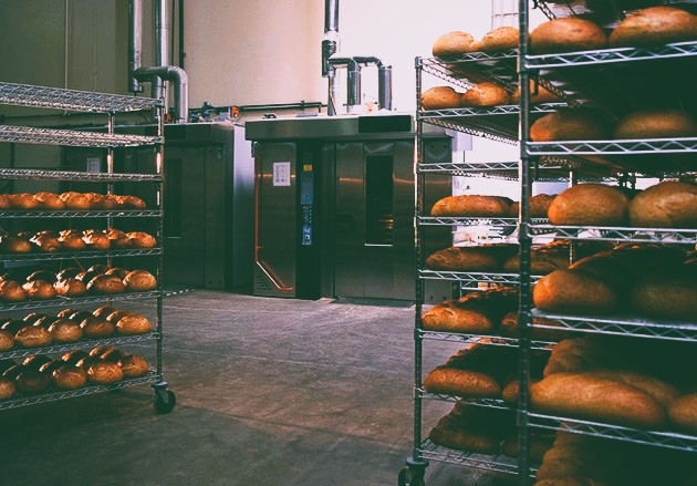 Freshly baked artisan bread loaves cooling on metal racks in a commercial bakery kitchen.