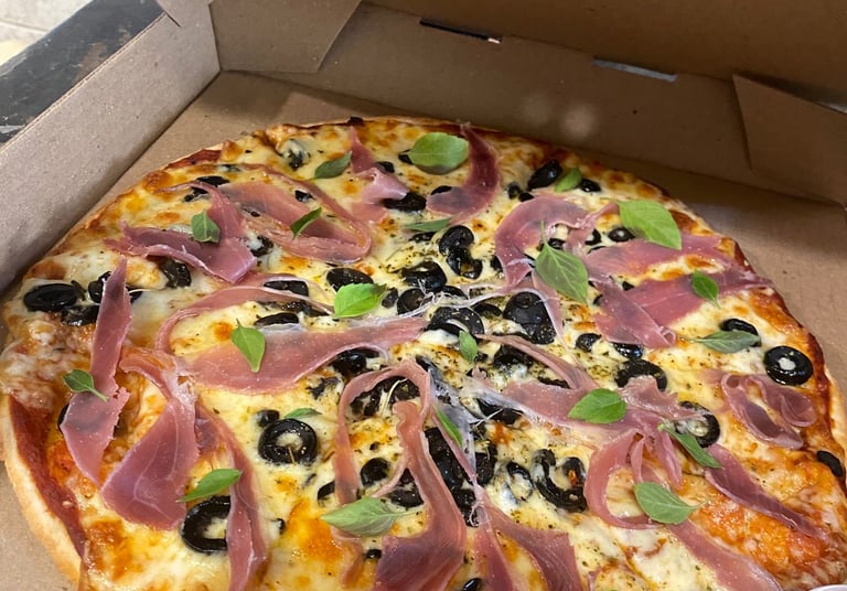 Pizza recién hecha para llevar en una caja de cartón, con jamon serrano, aceitunas negras y albahaca