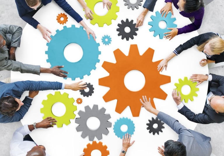 A group of people seated around a table are collaboratively arranging colorful gear-shaped pieces, symbolizing teamwork