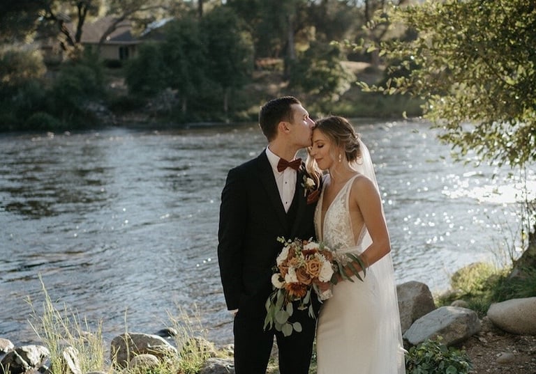 Groom kisses bride's forehead during an outdoor riverfront wedding ceremony with scenic nature views.