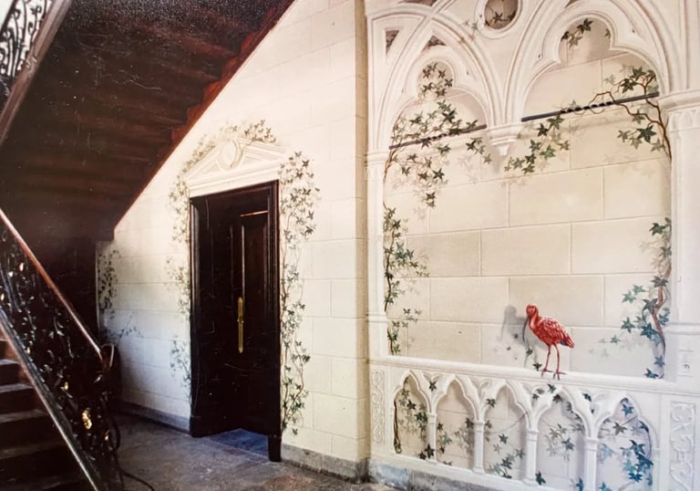 Hallway with wooden staircase and large mural of faux sandstone, ivy, gothic arches, and red bird.