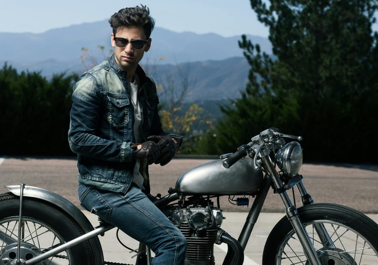 a man in a denim jean jacket and sunglasses sitting on a motorcycle in the mountains