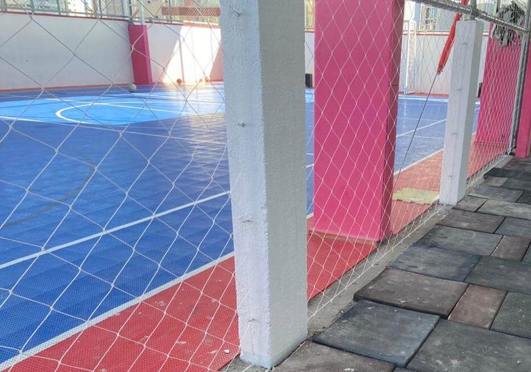 Lateral de quadra com piso azul e borda vermelha, cercada por rede de proteção.