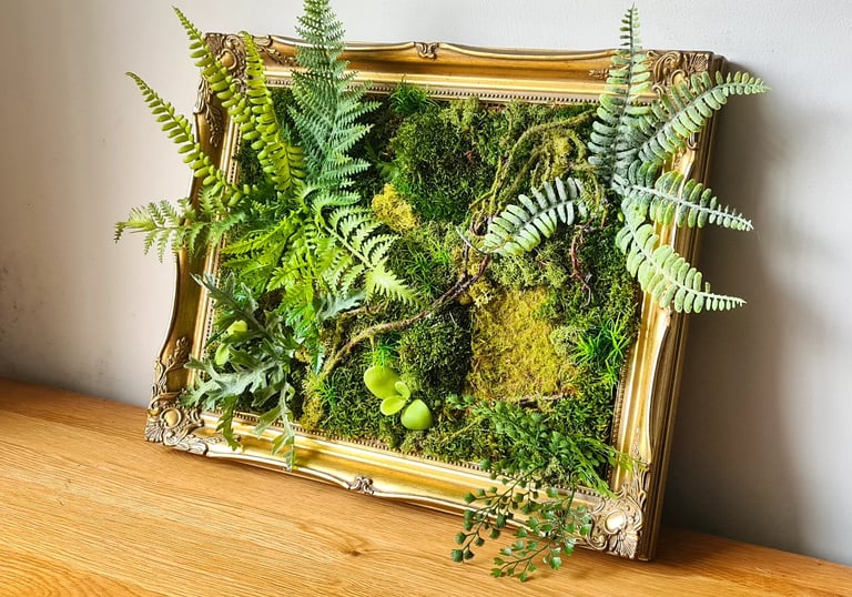 Preserved moss wall art with green ferns in an ornate gold vintage picture frame on a wooden table.