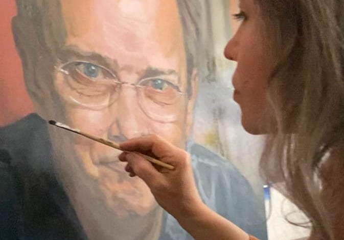 Close-up on Stefanie adding last details to oil portrait of smiling man.
