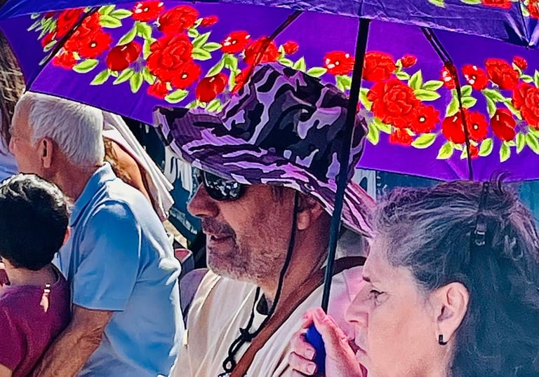 A man in a purple hat and a woman sit under a floral purple sun umbrella at an outdoor event.