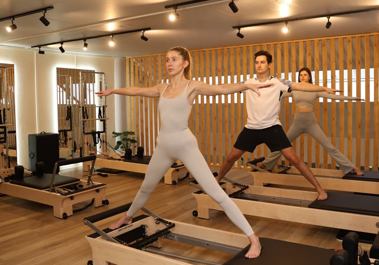Three people practicing Pilates Warrior II pose on reformers in a modern studio.