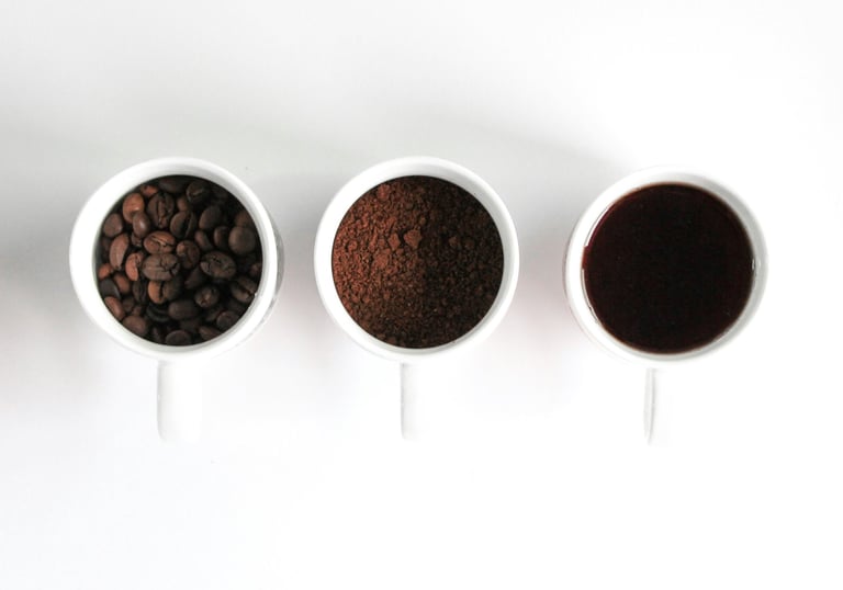 Three white cups in white background. one with coffee beans. one with coffee grinds. one with black coffee.