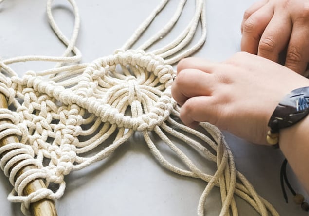 Manos elaborando un colgante de pared de macramé con cordón de algodón blanco sobre una madera.