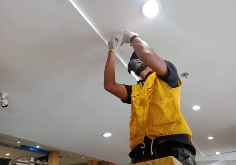 Electrician in a yellow safety vest installing recessed LED lighting in a white ceiling.
