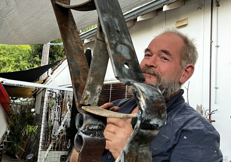 A bearded blacksmith holding a large custom metal sculpture with welded iron joints in an outdoor workshop.