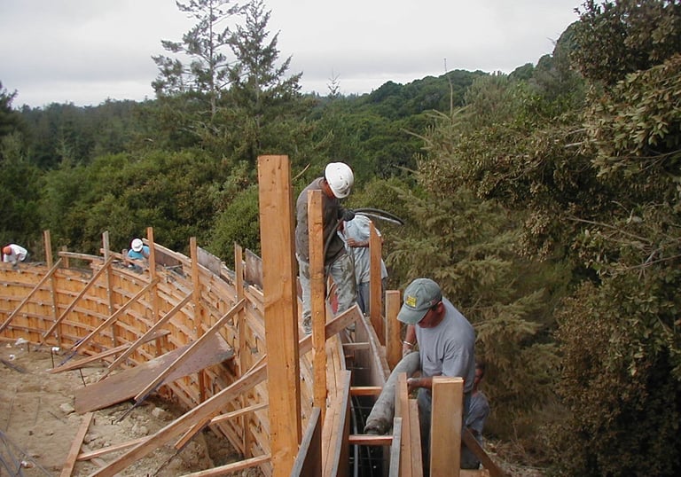 Construction workers pour concrete into a curved wooden retaining wall frame on a hillside.