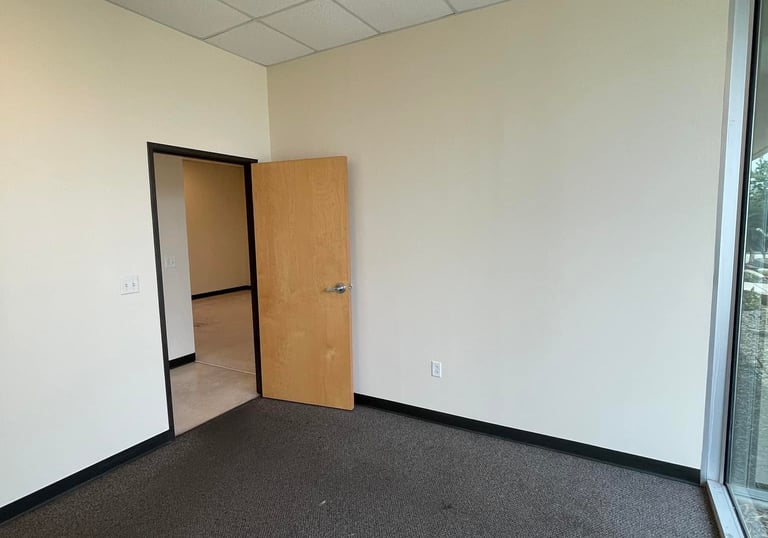 Empty commercial office space with grey carpet, beige walls, and a wooden door for rent.
