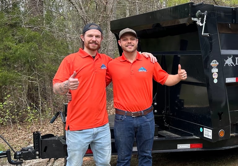 Two men in orange shirts giving thumbs up by a black dump trailer for  junk removal services.