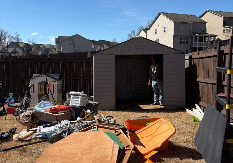 A man stands in an open backyard storage shed surrounded by cluttered outdoor items and lawn equipment.