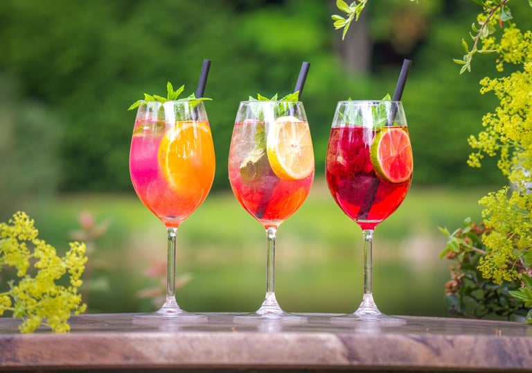 Drei lebendige Sommer-Cocktails in hohen Stielgläsern auf rustikalem Holz, garniert mit Früchte