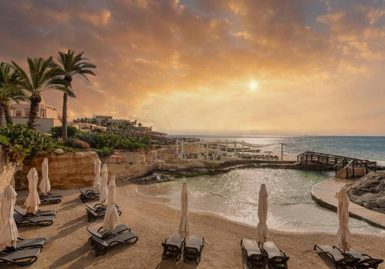 Luxury beach resort in Malta featuring palm trees and sun loungers at sunset by the Mediterranean Sea.