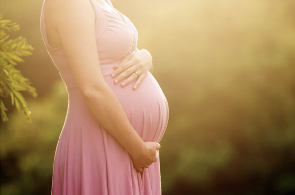 Pregnant woman in a pink maternity dress holding her baby bump outdoors in golden sunset light.
